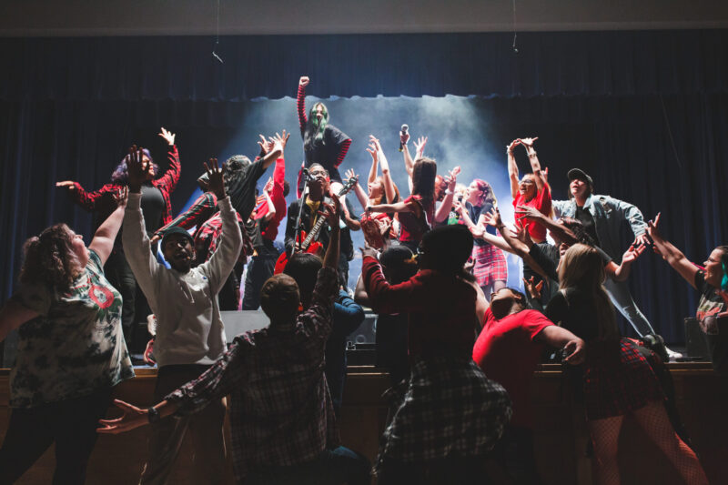 A diverse group of creative arts academy students on a stage performing with their arms raised toward a person playing an electric guitar.